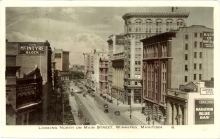 Black-and-white photograph with lightly coloured buildings in yellow and pink, depicting the view of a city street with brick buildings and storefronts. There are signs on three buildings for The McIntyre Block, Marathon Blue Gas, and Bank of Montreal