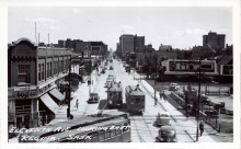 Photo en noir et blanc d'une scène d'une rue, prise en hauteur. Une large rue où circulent des piétons sur les trottoirs, il y a des vitrines de commerce de chaque côté, et trois tramways qui circulent au centre de la rue.