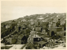 Photo en noir et blanc de petits bâtiments résidentiels dans un village ou une ville bâti sur une colline. Il y a de la neige au sol et il semble que ce soit l'hiver.