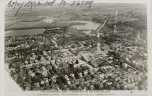 Black-and-white aerial photograph of a town with a river visible in the distance. Text has been handwritten in the top-left corner of the photograph.