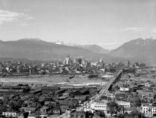 Black-and-white photograph of a bird's eye view of a city. Several buildings are pictured on either side of a river. A bridge connecting the two banks is pictured on the right-hand side of the photo. Several mountain peaks are visible in the background.