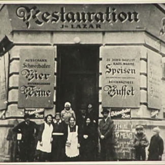 Black-and-white grainy photograph of a group of about 8 people standing in front of the entrance to a restaurant. The building has large signs on either side of the door, written in German. A couple young children look at the group from the side.