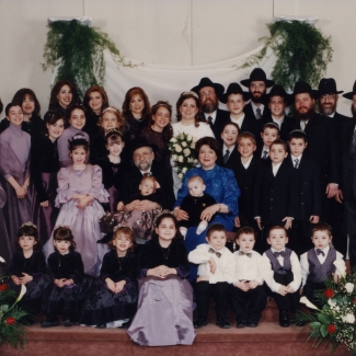 Photo en couleur d'un grand groupe d'environ 30 personnes rassemblés pour une photo de groupe de trois ou quatre rangées, certains sont assis dans la première rangée. Ils sont tous habillés de manière formelle. Les femmes portent des robes et les hommes des complets et des chapeaux. Il y a de grands bouquets de fleurs de chaque côté du groupe.