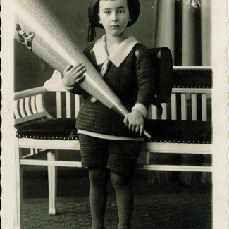 Black-and-white studio portrait photograph of a young boy wearing a school uniform and hat, looking at the camera in a pensive pose. He holds a large wooden cone-shaped object. There is white writing in German at the bottom of the image, and the edges of the photograph are slightly worn.