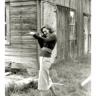 Black-and-white photograph of a woman standing in front of a barn. She holds up a rifle and aims it towards the left of the camera.