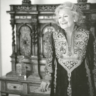 Black-and-white photograph of an elderly woman dressed in an embroidered tunic, leaning against an elaborately carved wooden hutch.