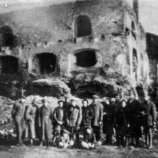 Photo en noir et blanc d'un groupe de 15 personnes se tenant ensemble en ligne à l'extérieur, deux enfants sont assis sur le sol, devant les ruines d'un bâtiment. Le bâtiment n'a plus de toit et plusieurs cadres de fenêtres sont sans fenêtres.