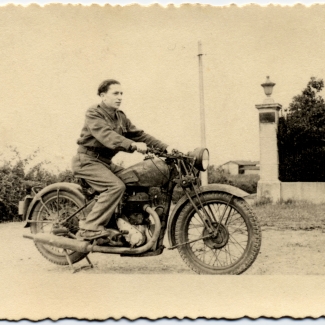 Photo en noir et blanc d'un jeune homme sur une motocyclette, regardant vers la droite de la caméra. Il y a une haie derrière lui et un bâtiment en arrière-plan.