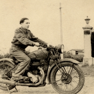 Black-and-white photograph of a young man posing on the back of a motorcycle, looking forwards towards the right of the camera. There is a hedge behind him and a building in the distant background.