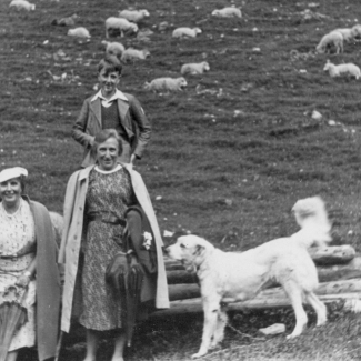 Photo en noir et blanc d'un jeune homme debout derrière un chien blanc et de deux femmes, l'une d'elles est assise sur une pile de bûches. Un troupeau de moutons se promène derrière eux.