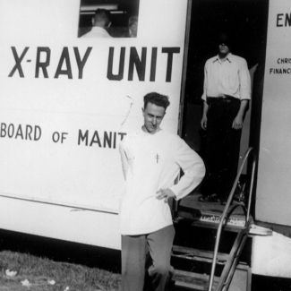 Photo en noir et blanc d'un homme debout, la main appuyée sur la hanche gauche, devant un grand camion blanc sur lequel il est inscrit “X-RAY UNIT” sur un côté. Un homme est debout derrière lu dans l'entrée du camion, son visage est caché par un ombrage.