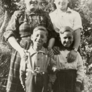 Black-and-white photograph of a two women standing behind two young twin boys, all 4 of them smiling at the camera. They are standing outside and in what looks like summer weather, due to the women’s rolled-up sleeves, and the boys’ shorts.