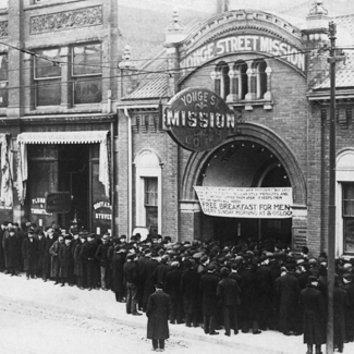 Black-and-white photograph of an outdoor street scene. A group of people are lined up outside a building.