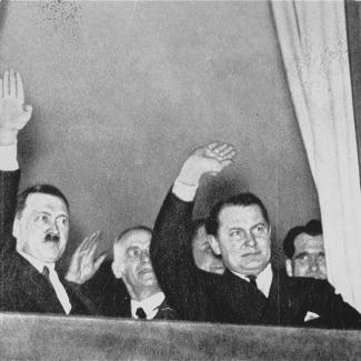 Black-and-white photograph of two men in waving out a window. The photograph is taken from below, outside. The man on the left has a moustache, and there are men behind them in the background.