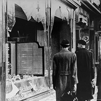 Black-and-white photograph of three men in hats and jackets, standing on a sidewalk and looking at two shattered storefront windows. Two men have their back to the camera.