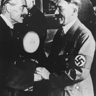 Photo en noir et blanc de deux hommes souriant et se serrant la main. Les deux hommes portent une moustache, et un des deux hommes porte un complet plus pâle avec un brassard avec une swastika.