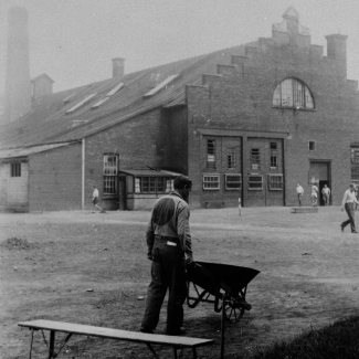 Black-and-white photograph of a man, with his back to the camera, pushing a wheelbarrow in a yard. There is a bench in the foreground, and a building in the far background. The man wears a uniform with a big circle on his back.