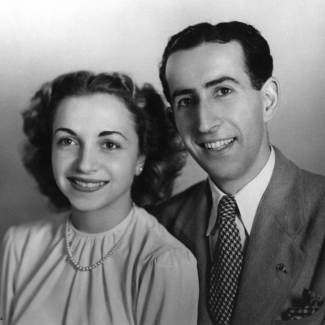 Black-and-white studio photograph of a man and woman sitting together and smiling at the camera. The man wears a suit, and the woman wears a blouse with a pearl necklace. Both have dark brown hair.