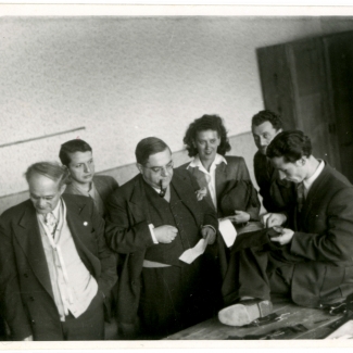 Photo en noir et blanc d'un groupe de six personnes, cinq hommes et une femme, se tenant debout dans une pièce. Un homme fume la pipe et observe un autre homme assis à une table qui cout un tissus à la main.