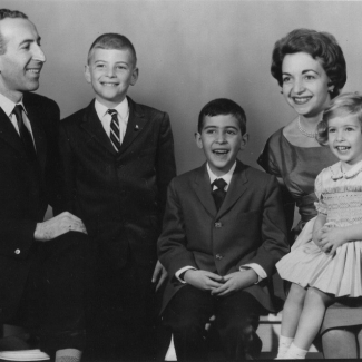 Photo en noir et blanc prise en studio d'un homme et d'une femme avec leur trois jeunes enfants, souriant à la caméra. L'homme et les deux garçons portent des complets, et la femme et sa fille portent des robes.