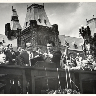 Black-and-white photograph of a man in a suit speaking into two microphones at a podium outdoors. He is surrounded by a group of people, some sitting, with a large building in the background.