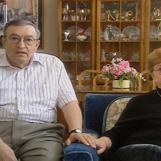 Colour photograph of a elderly man and woman sitting together indoors, holding hands. The woman sits on a blue arm chair. There is a display cabinet containing dishes and chinaware in the background.