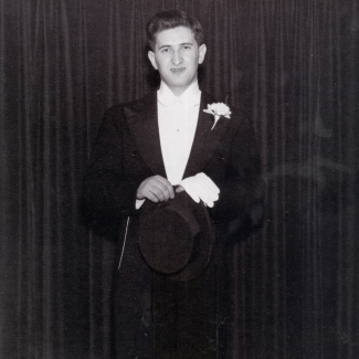 Photo en noir et blanc d'un jeune homme portant un tuxedo , se tenant devant un rideau de couleur foncée.