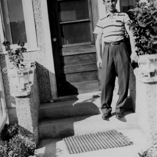 Black-and-white photograph of a young man standing outdoors, on the first step of an entrance to a house. He wears a short-sleeved striped shirt and trousers.