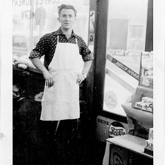 Photo en noir et blanc d'un homme se tenant à l'intérieur d'une épicerie. Il a les mains appuyées sur ses hanches et il porte un tablier blanc, il est debout près de la porte d'entrée du commerce. Il y a les mots “LYONS CONFECTIONARY STORE” écrits à l'envers dans la fenêtre derrière lui.