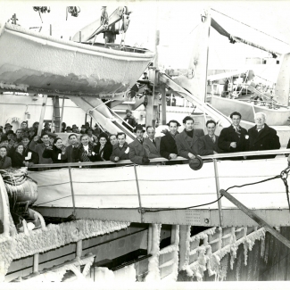 Photo en noir et blanc d'un groupe de 15 personnes, portant manteaux et chapeaux, souriant et saluant la caméra depuis la passerelle d'un navire. Plusieurs autres personnes attendent leur tour pour débarquer du navire. Un bateau de sauvetage dont les côtés sont glacés est suspendu dans le coin supérieur gauche.