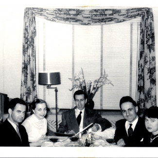 Black-and-white photograph of a group of 5 adults casually sitting at a dining table with glasses of wine, smiling at the camera. The men wear suits.