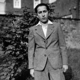Black-and-white photograph of a young man standing outdoors in what appears to be a backyard, with a fence and shrub behind him. He wears a suit jacket and collared shirt, with his hair combed back.