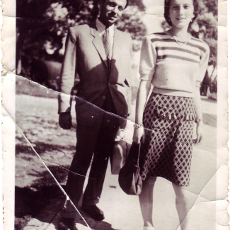 Black-and-white photograph of a man and woman standing together outdoors on the pavement, with trees in the background. The man wears a suit, and the woman wears a blouse and polka-dot skirt.