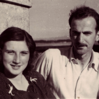 Black-and-white photograph of a man and woman, sitting together outdoors. The woman has shoulder-length brown hair, and the man has a moustache, wearing a light collared shirt.