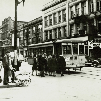 Photo en noir et blanc d'une scène d'une, prise au niveau de la rue. Un groupe de personnes attendent pour embarquer dans un tramway stationné dans la rue.