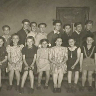 Black-and-white photograph of a classroom, grouped together in two rows of about 10-12 children each. The girls wear dresses, sitting in the front row and the boys stand in the back with collared shirts.