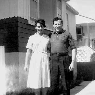 Black-and-white photograph of a man and woman smiling and standing outside the entrance to a house. It appears to be summer. The woman wears a short-sleeved dress.