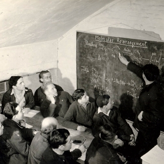 Photo en noir et blanc d'un groupe d'environ douze hommes regardant un tableau. Un des hommes est debout avec un morceau de craie et pointe vers une équation écrite sur le tableau. Les autres hommes sont assis à la table, certains prennent des notes avec papier et crayon.