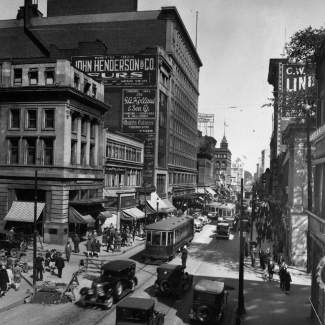 Photographie en noir et blanc d'une scène d'une rue achalandée où circulent des voitures d'époque et des tramways. Des piétons marchent dans chaque direction sur des trottoirs et il y a des panneaux d'affichage publicitaire sur les murs extérieurs des édifices.