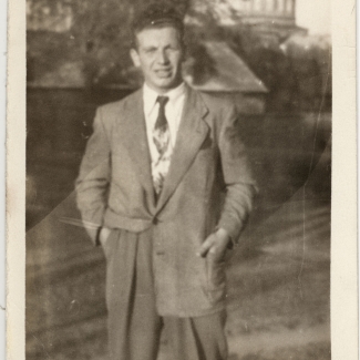 Black-and-white photograph of a man wearing a suit and tie, standing outdoors on a field. A building tower is visible in the background.