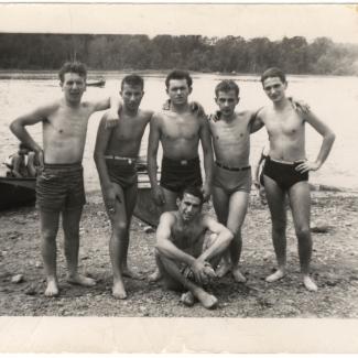 Black-and-white photograph of a group of 6 teenage boys standing arm-in-arm on a beach, with water in the background. The young men wear bathing suits, and one of them sits on the ground.