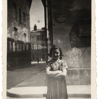 Black-and-white photograph of a young girl standing a street with her arms crossed, smiling at the camera. She stands near the corner of the street, with the road and sidewalk continuing in the background.