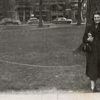 Black-and-white photograph of a woman smiling and leaning against a street light in front of a large field with buildings and cars in the background. She holds a purse and book in her hand.