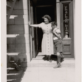 Black-and-white photograph of a woman and a boy standing at the doorstep of a ground floor apartment. The woman has her arm around the son and rests her other arm against the entrance to the apartment.