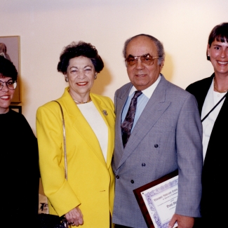 Colour photograph of an elderly man and woman standing between two other women. The group of four smile at the camera. The man wears a suit and holds a framed document.