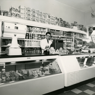 Photo en noir et blanc d'un homme se tenant derrière une caisse enregistreuse dans une grand épicerie. Il se tient devant une étagère remplie de produits et il y a un grand comptoir de présentation devant lui.