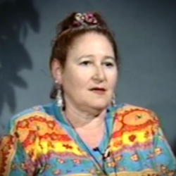 Screenshot of Holocaust survivor Helene Goldflus video testimony. She is sitting in front of a blue wall with a plant in the background, and looking to the right of the camera. The camera shows her face and shoulders.