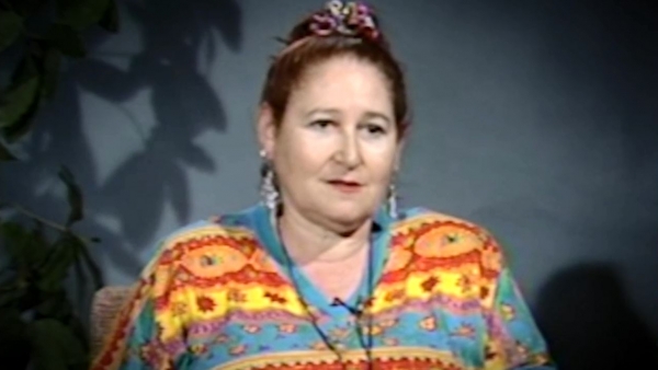 Screenshot of Holocaust survivor Helene Goldflus video testimony. She is sitting in front of a blue wall with a plant in the background, and looking to the right of the camera. The camera shows her face and shoulders.