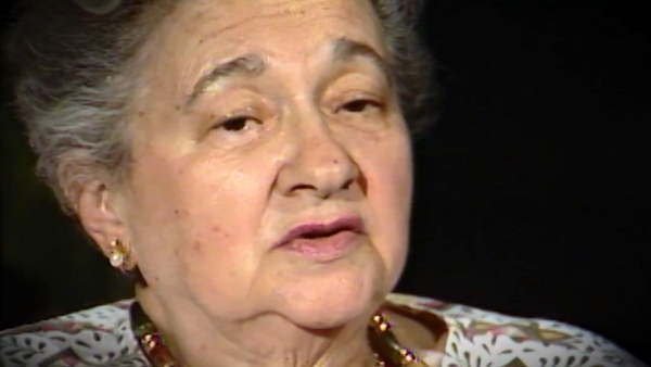 Screenshot of Holocaust survivor Mania Kay video testimony. She is sitting in front of a black background, and looking to the right of the camera. The camera shows her face and shoulders.